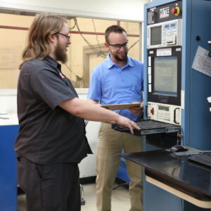 Two engineers working on a high-voltage testing system, with one operating the equipment and the other reviewing data on a clipboard in a laboratory setting.